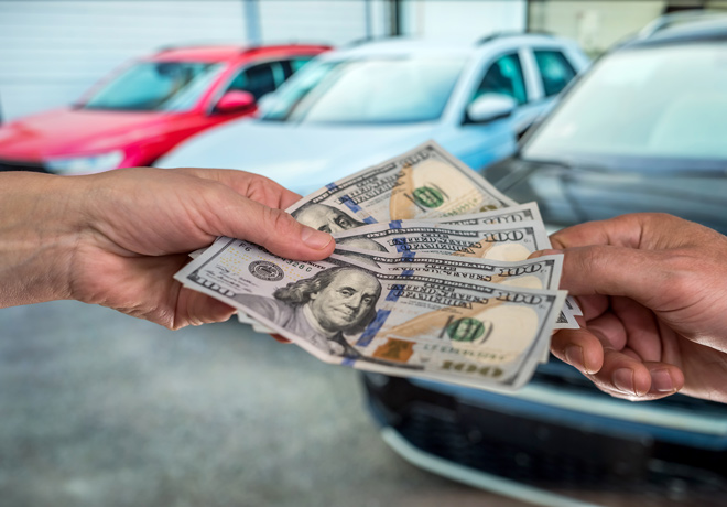 A man's hand paying several hundred dollars in cash to another hand with cars in the background.